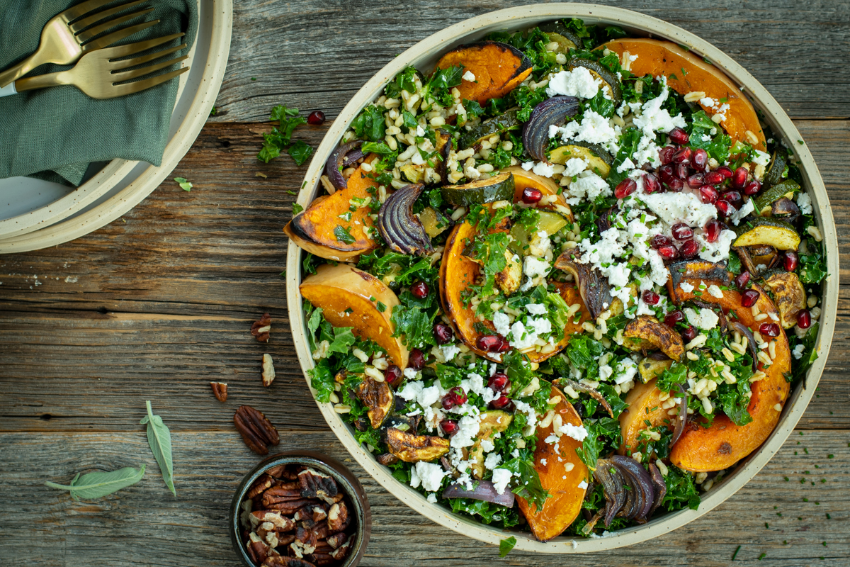 Bovenaanzicht van een ronde schaal gevuld met een salade van geroosterde pompoen met kamut, boerenkool en feta; op een houten ondergrond.