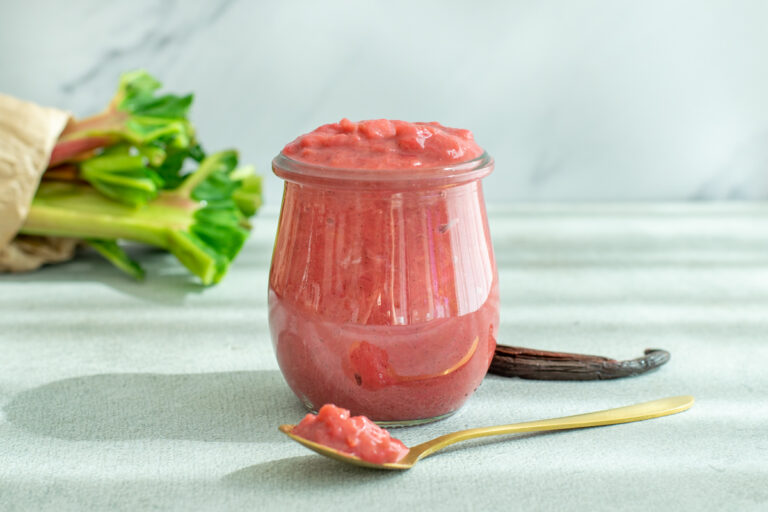 Een potje rode rabarbercompote, met vanillezaadjes, op een lichtgrijze achtergrond.