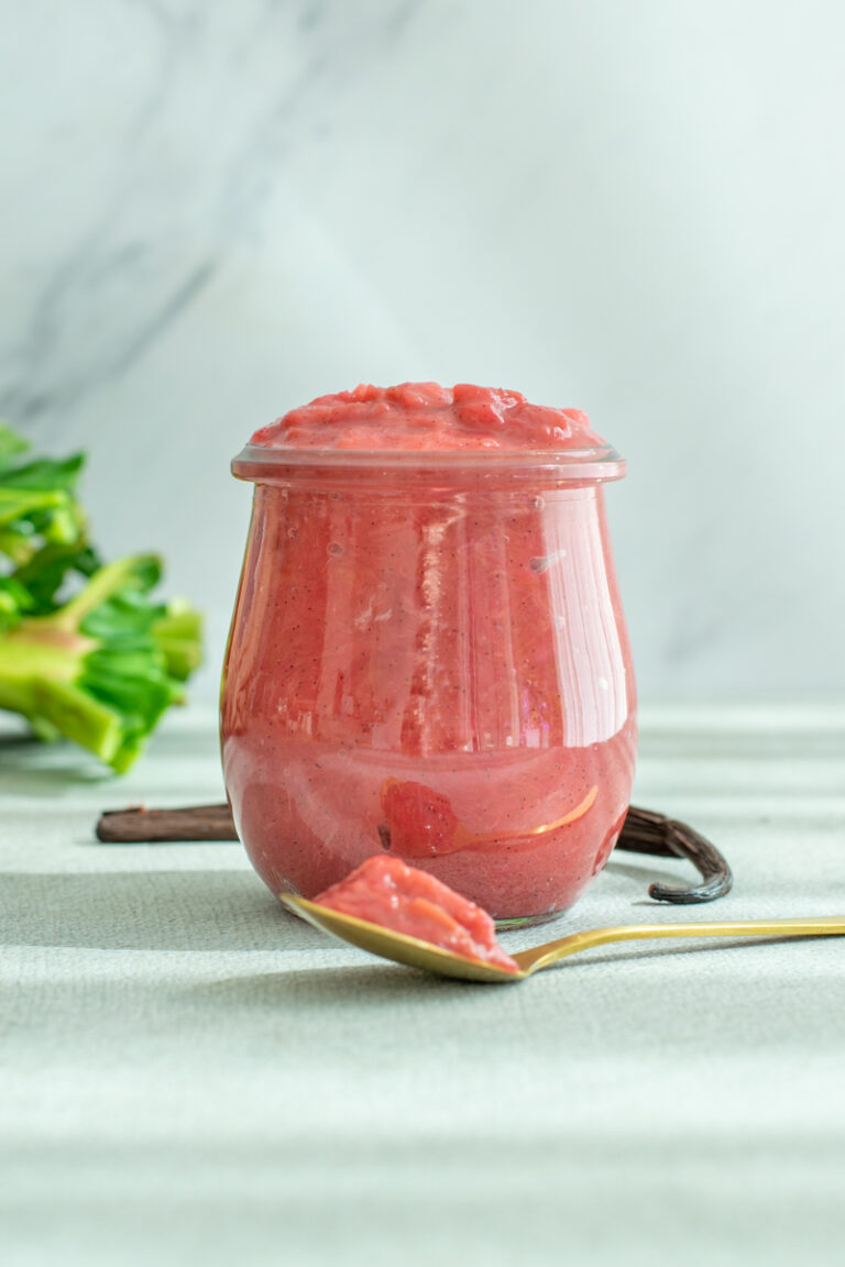 Een potje rode rabarbercompote, met vanillezaadjes, op een lichtgrijze achtergrond.