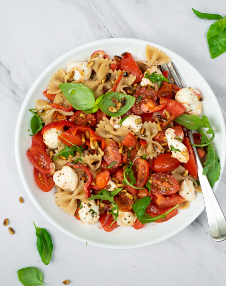 Bovenaanzicht van een pastasalade caprese, met farfalle, bolletjes mozzarella en verschillende soorten tomaat, afgewerkt met vers basilicum, op een lichtegrijze ondergrond.