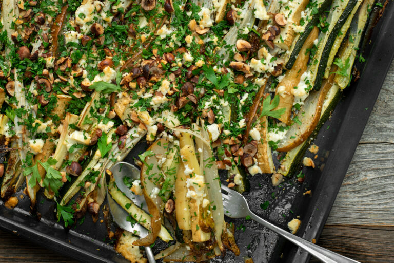 Bovenaanzicht van een bakplaat met geroosterde witloof, pastinaak en peer, afgewerkt met groene kruiden, hazelnoten en feta.