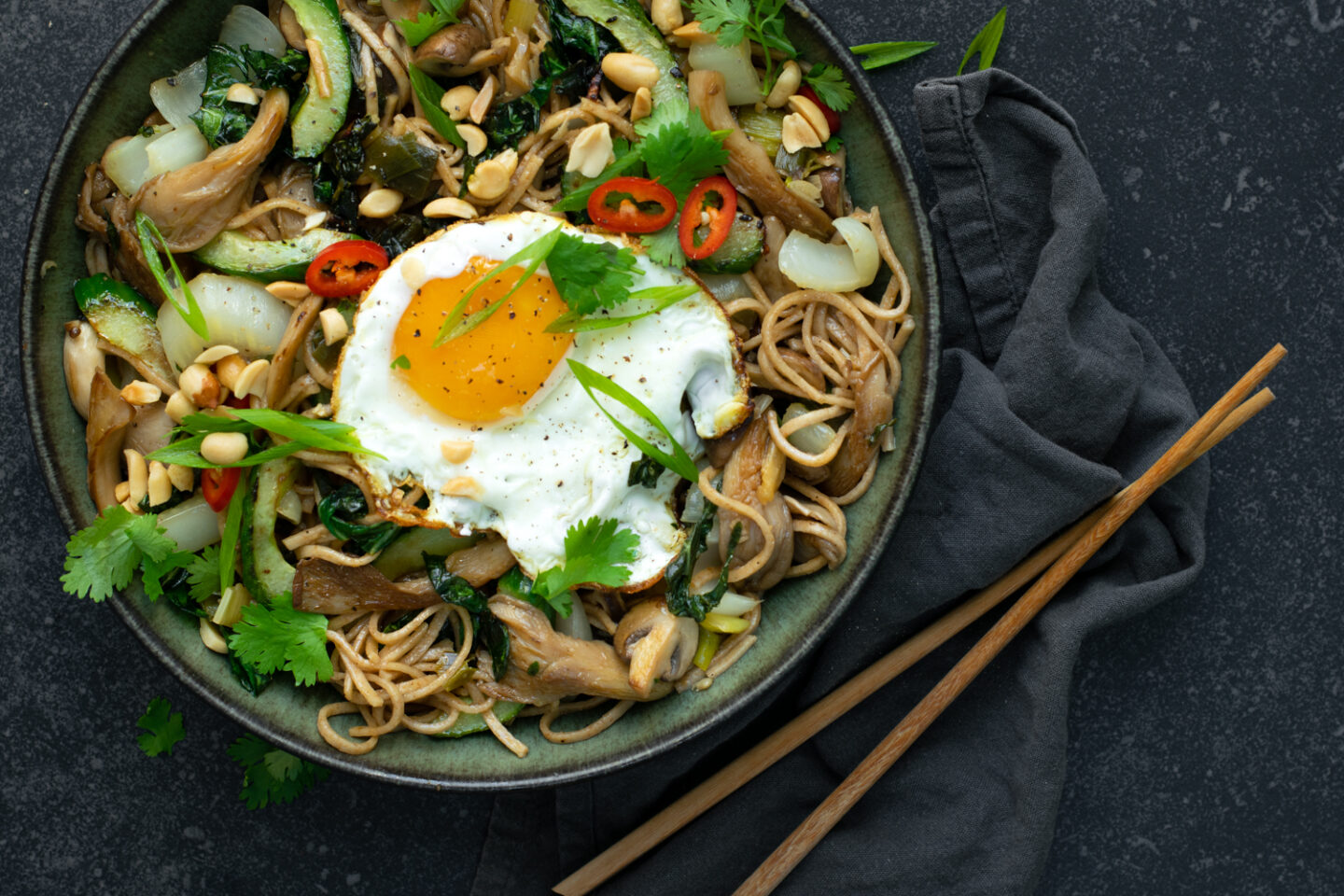 Een donker bord met noedels met champignons, paksoi en een spiegelei, op een donkere achtergrond.