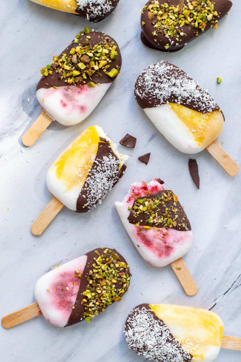 Bovenaanzicht van 8 kokos-chocolade ijslolly's. De ijsjes zijn wit-geel of wit-roze gemarmerd en half bedekt met een laagje chocolade, afgewerkt met pistachenoten of kokosrasp.