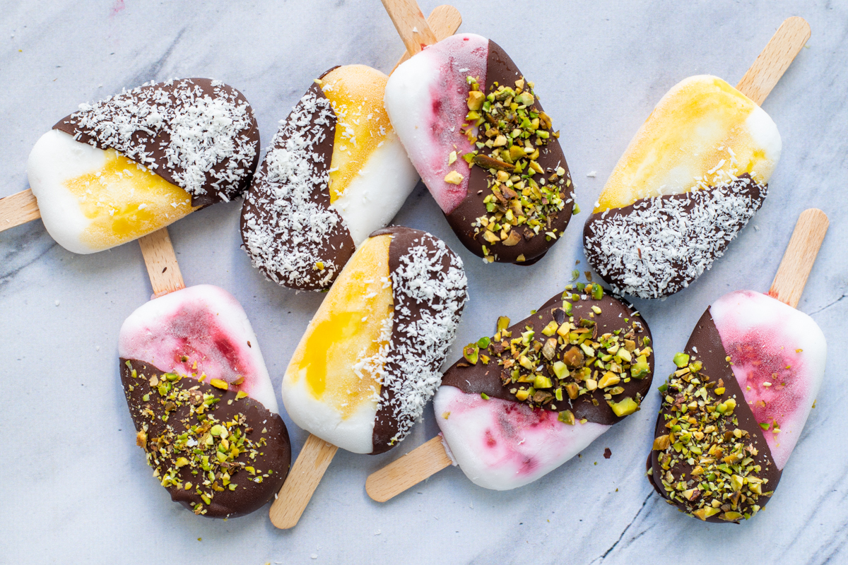 Bovenaanzicht van 8 kokos-chocolade ijslolly's. De ijsjes zijn wit-geel of wit-roze gemarmerd en half bedekt met een laagje chocolade, afgewerkt met pistachenoten of kokosrasp.