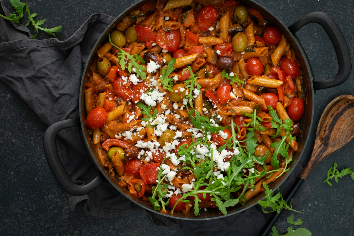 Bovenaanzicht van een ronde zwarte stoofpan op een zwarte ondergrond. In de pan zit penne met een tomatensaus, kerstomaatjes en olijven, afgewerkt met feta en rucola.