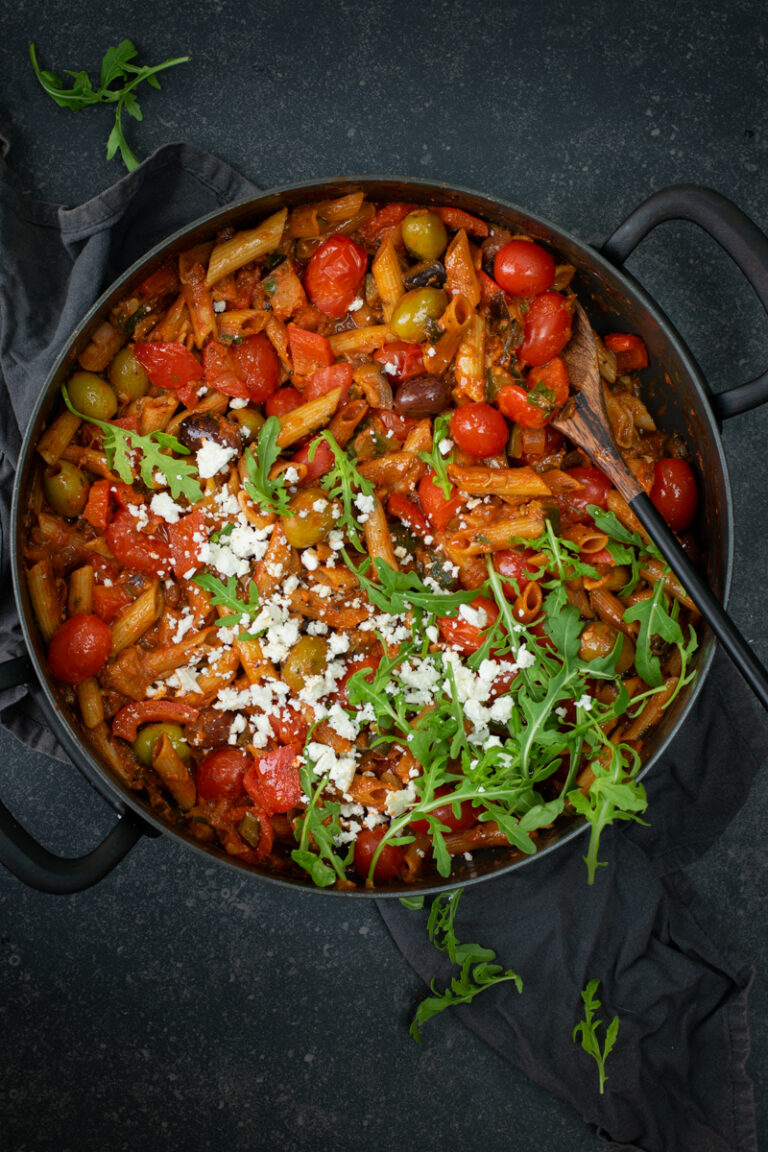 Bovenaanzicht van een ronde zwarte stoofpan op een zwarte ondergrond. In de pan zit penne met een tomatensaus, kerstomaatjes en olijven, afgewerkt met feta en rucola.