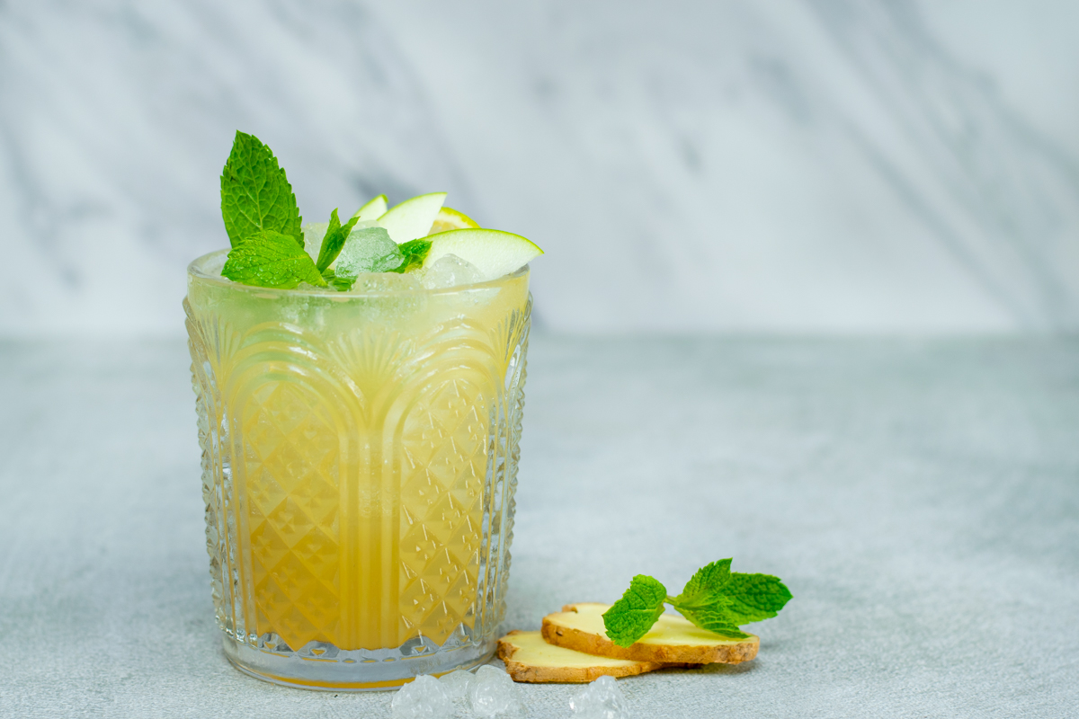 Zijaanzicht van een glas gevuld met appel-gember mocktail, afgewerkt met munt en schijfjes appel. Tegen een lichtgrijze achtergrond.