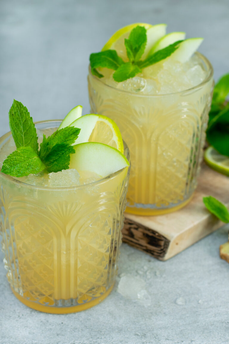Zijaanzicht van twee glazen gevuld met appel-gember mocktail, afgewerkt met munt en schijfjes appel. Tegen een lichtgrijze achtergrond.