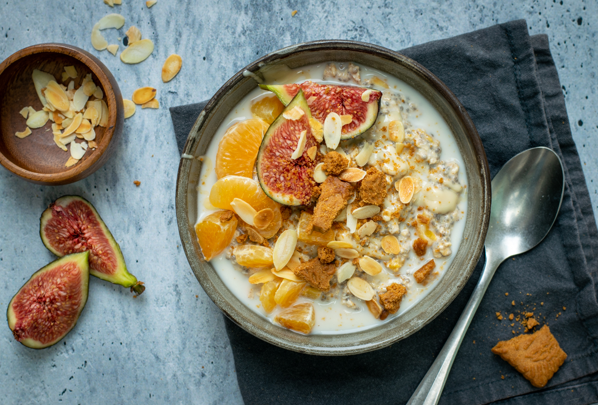 Bovenaanzicht van een donkergrijze kom met speculaas overnight oats, afgewerkt met mandarijn, vijg, amandelschilfers en speculaaskruimels. Op een lichtgrijze, beton ondergrond.