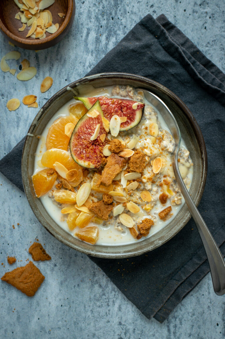Bovenaanzicht van een donkergrijze kom met speculaas overnight oats, afgewerkt met mandarijn, vijg, amandelschilfers en speculaaskruimels. Op een lichtgrijze, beton ondergrond.