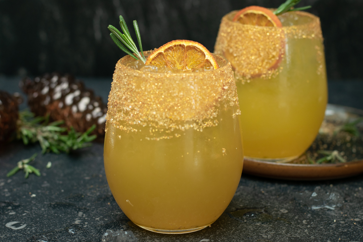Twee bolle glazen met een goudkleurige chai-ginger mocktail, afgewerkt met sinaas en rozemarijn, op een donkere achtergrond.