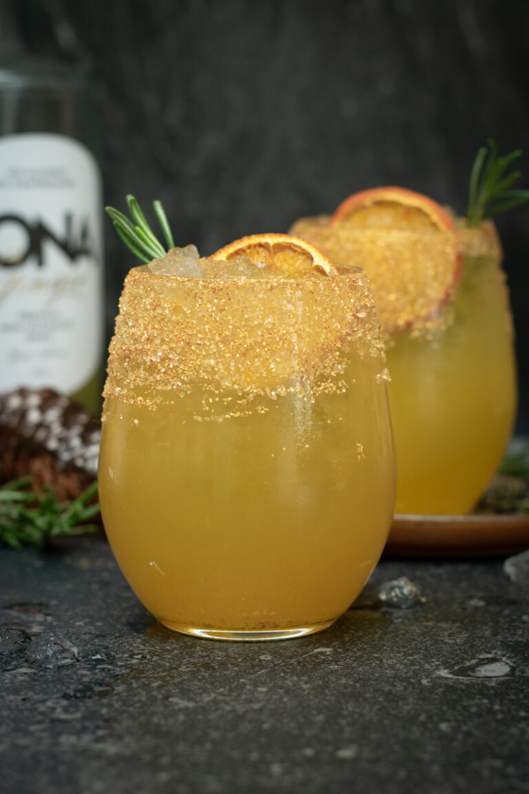 Twee bolle glazen met een goudkleurige chai-ginger mocktail, afgewerkt met sinaas en rozemarijn, op een donkere achtergrond.