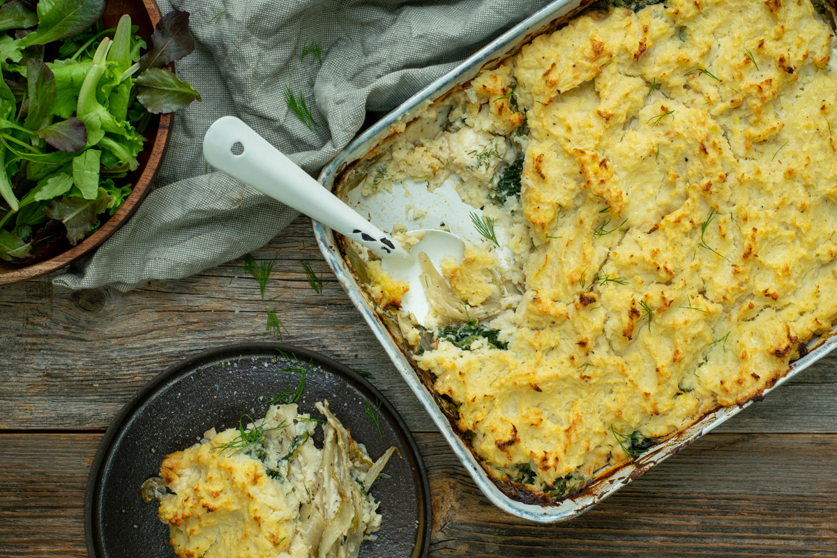 Bovenaanzicht van een aangeschepte rechthoekige ovenschotel met een toplaag van gegratineerde puree. De vulling bestaat uit witloof, kaassaus en spinazie.