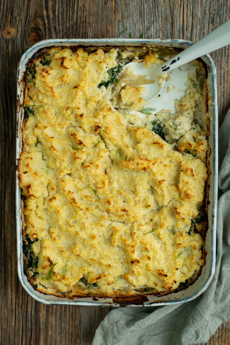 Bovenaanzicht van een aangeschepte rechthoekige ovenschotel met een toplaag van gegratineerde puree. De vulling bestaat uit witloof, kaassaus en spinazie.
