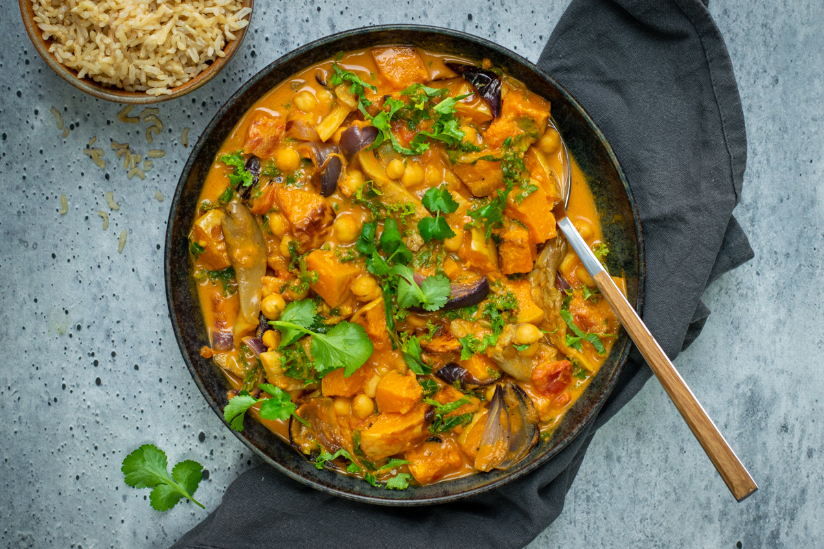 Bovenaanzicht van een zwarte kom met een oranje curry met pompoen en kikkererwten, op een grijze ondergrond, afgewerkt met verse koriander.