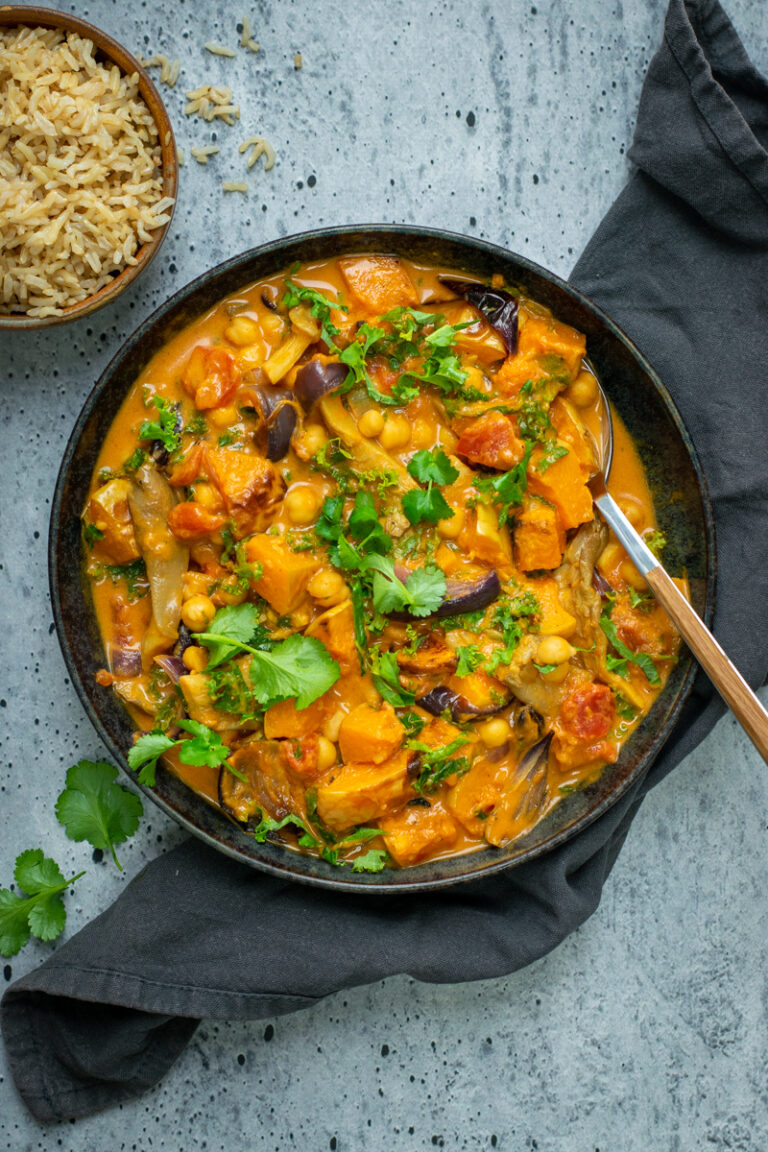 Bovenaanzicht van een zwarte kom met een oranje curry met pompoen en kikkererwten, op een grijze ondergrond, afgewerkt met verse koriander.