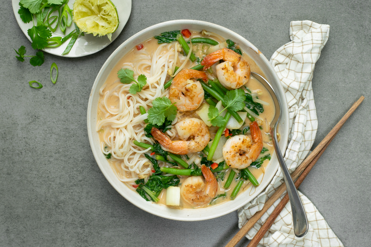 Bovenaanzicht van een wit diep bord op een grijze achtergrond. Het bord is gevuld met een noedelsoep met scampi's en groene groenten, afgewerkt met lente-ui en koriander.