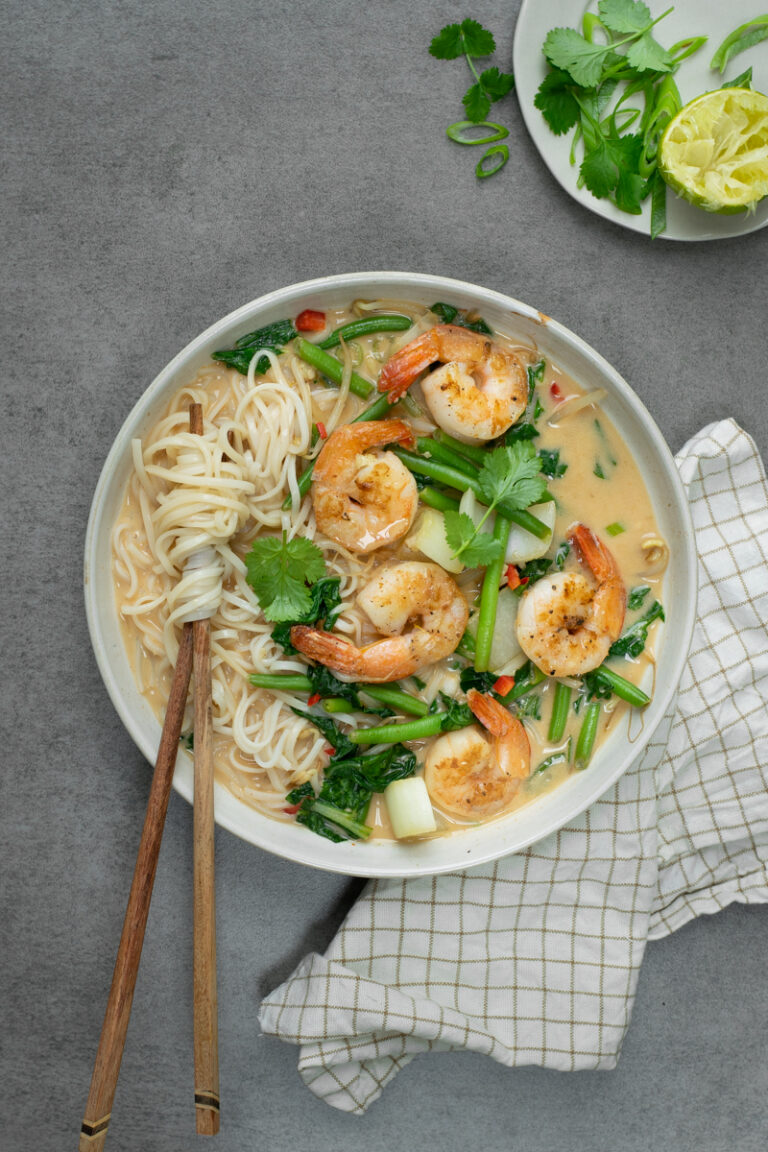 Bovenaanzicht van een wit diep bord op een grijze achtergrond. Het bord is gevuld met een noedelsoep met scampi's en groene groenten, afgewerkt met lente-ui en koriander.