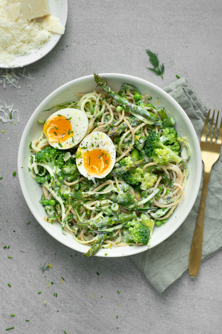 Bovenaanzicht van een spaghetti primavera met groene asperges, broccoli en erwtjes, afgewerkt met een gehalveerd zacht eitje.