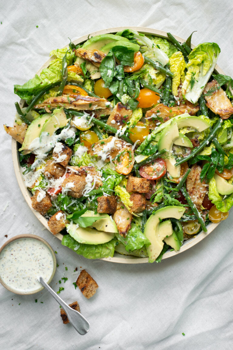 Bovenaanzicht van een Ceasar salade met extra avocado, prinsessenboontjes en tomaatjes. Er staat een potje dressing naast de salade.