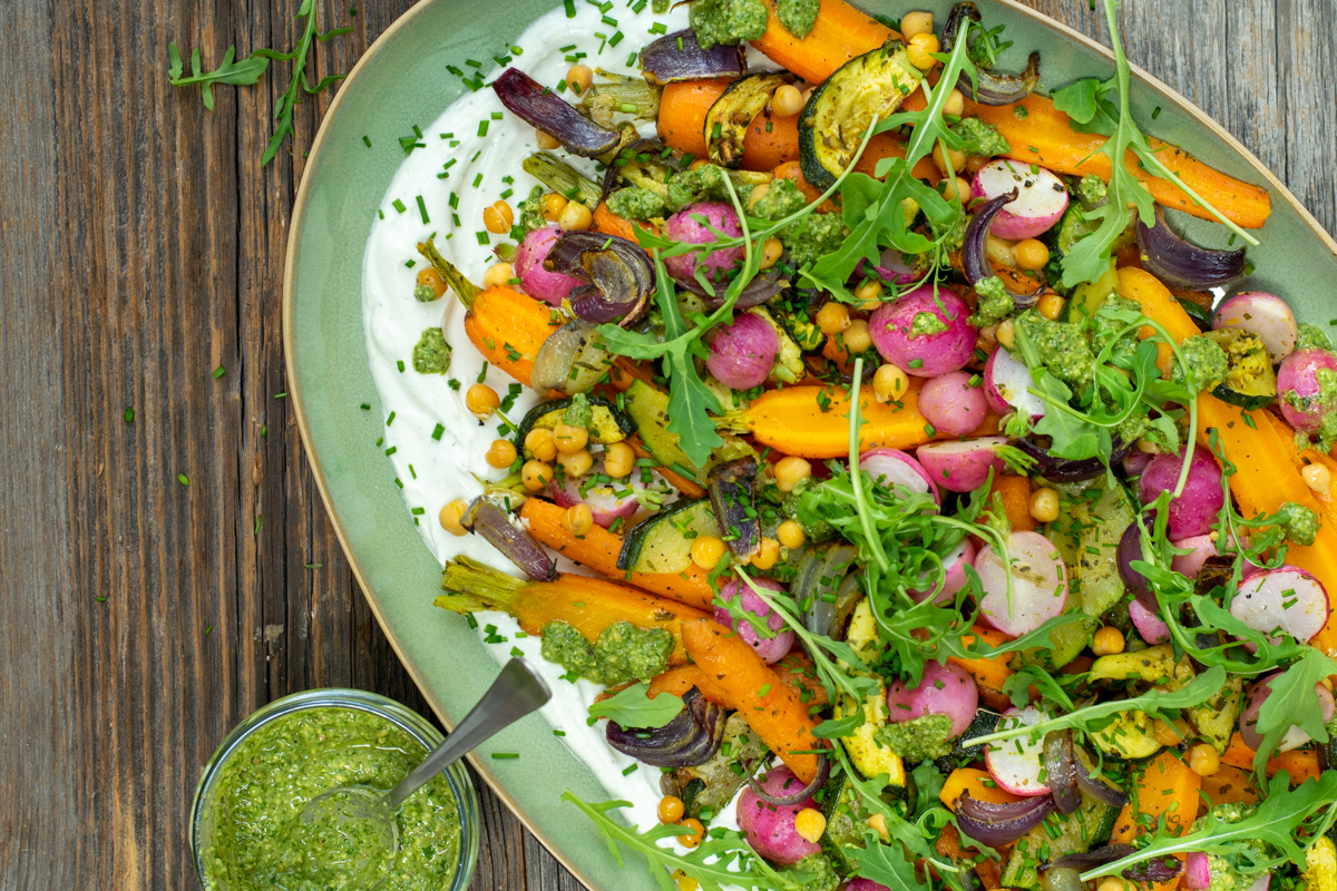 Bovenaanzicht van een azuurgroene, ovale schaal gevuld met cottagechees, geroosterde wortel, radijs, courgette en kikkererwten, afgewerkt met pesto en rucola