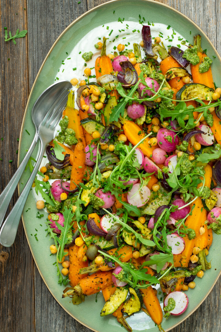 Bovenaanzicht van een azuurgroene, ovale schaal gevuld met cottagechees, geroosterde wortel, radijs, courgette en kikkererwten, afgewerkt met pesto en rucola