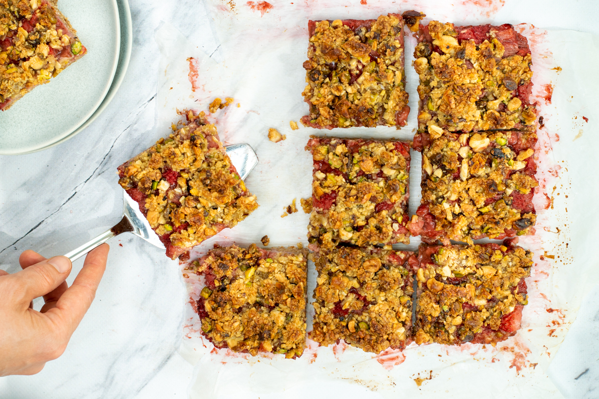 Bovenaanzicht van aardbei-crumble gebak, in vierkantjes gesneden, op een witte marmeren ondergrond, met een zilveren taartschep.