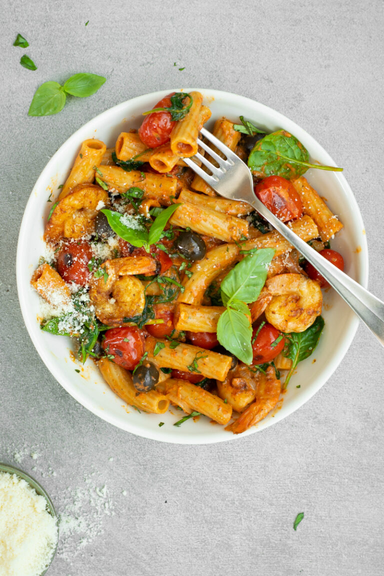 Bovenaanzicht van een diep wit bord met tortiglioni met paprikasaus, kerstomaten, zwarte olijven en scampi's, afgewerkt met basilicumblaadjes en Parmezaanse kaas.