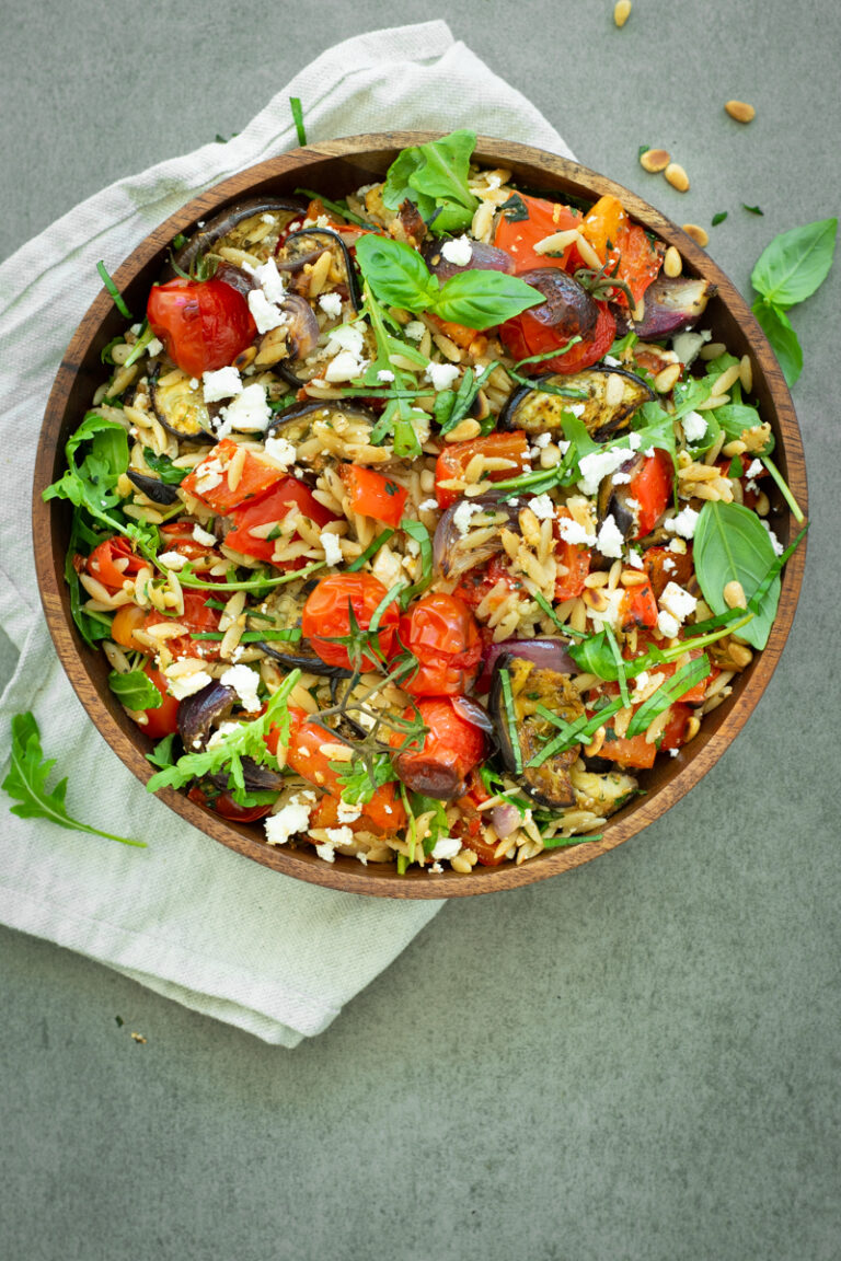 Bovenaanzicht van orzosalade met geroosterde aubergine, paprika en rode ui, afgewerkt met rucola, vers basilicum en feta.