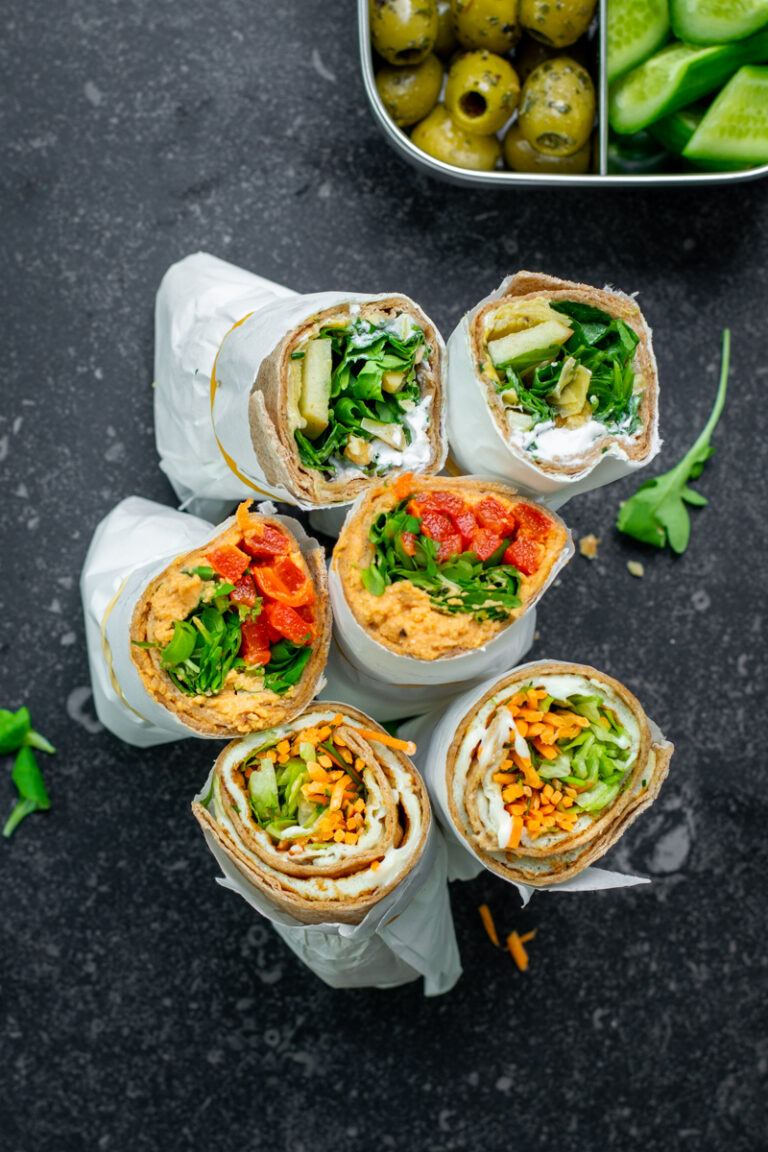 Bovenaanzicht van 3 opengesneden wraps, verpakt in wit boterhampapier, op een donkergrijze achtergrond.