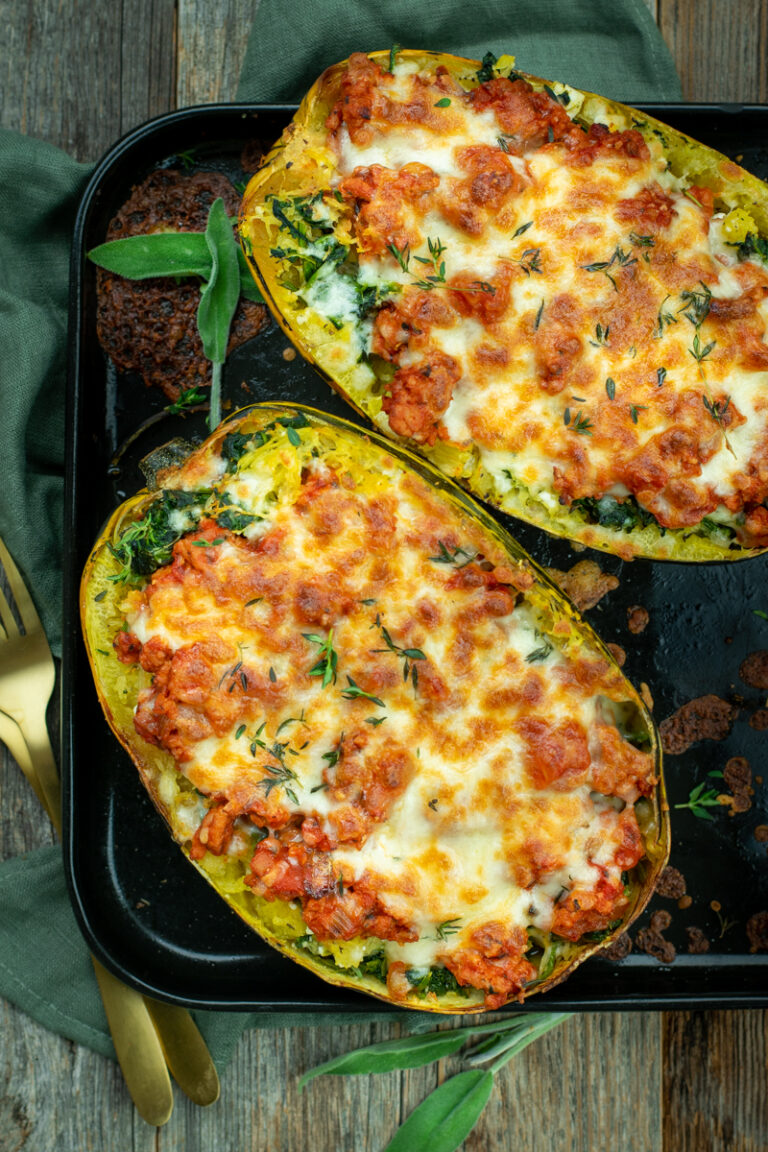 Bovenaanzicht van 2 halve spaghettipompoenen, gevuld met spinazie, bolognesesaus en gegratineerd met kaas. Op een zwarte bakplaat, op een houten ondergrond.