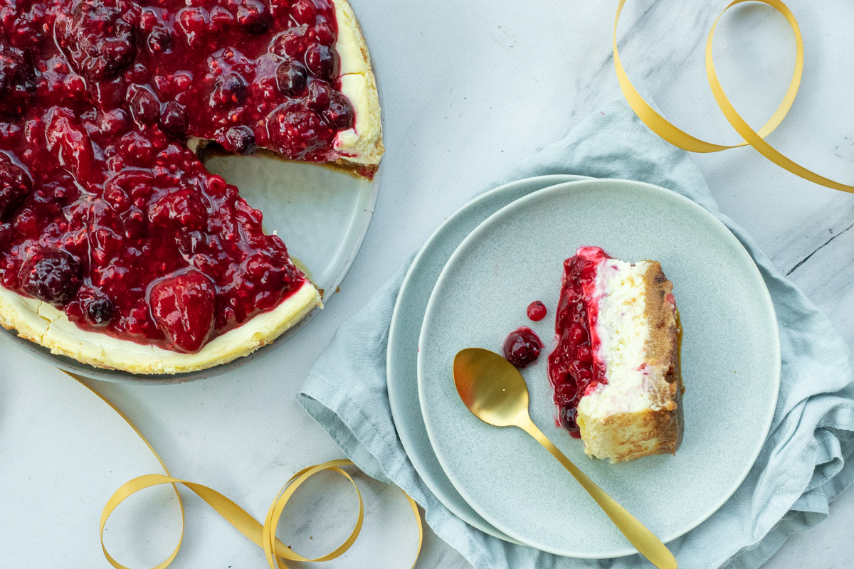 Bovenaanzicht van een ronde cheesecake met een toplaag van gestoofde rode vruchten en een bodem van speculaas. Er is één punt uitgesneden, die ligt op een bordje ernaast, met een gouden lepeltje erbij.