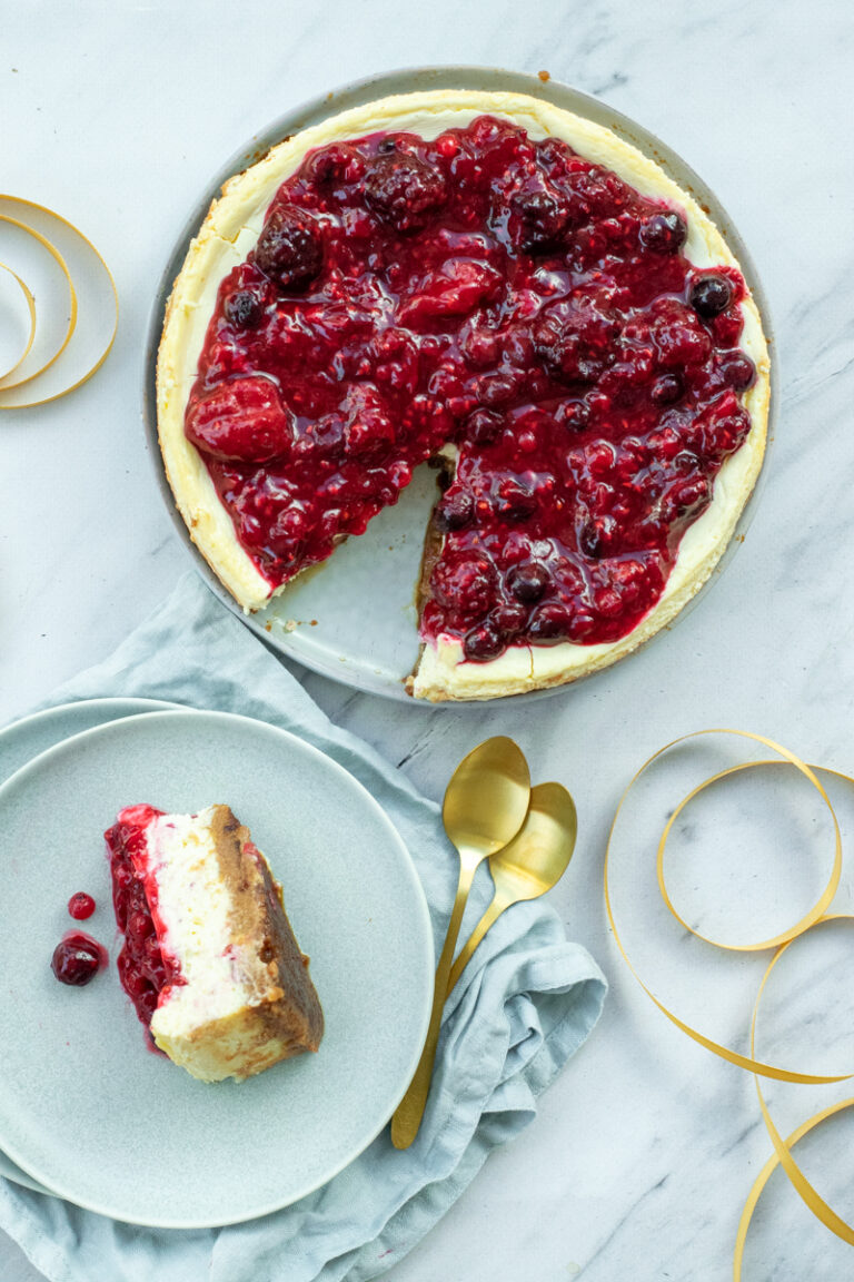 Bovenaanzicht van een ronde cheesecake met een toplaag van gestoofde rode vruchten en een bodem van speculaas. Er is één punt uitgesneden, die ligt op een bordje ernaast, met een gouden lepeltje erbij.