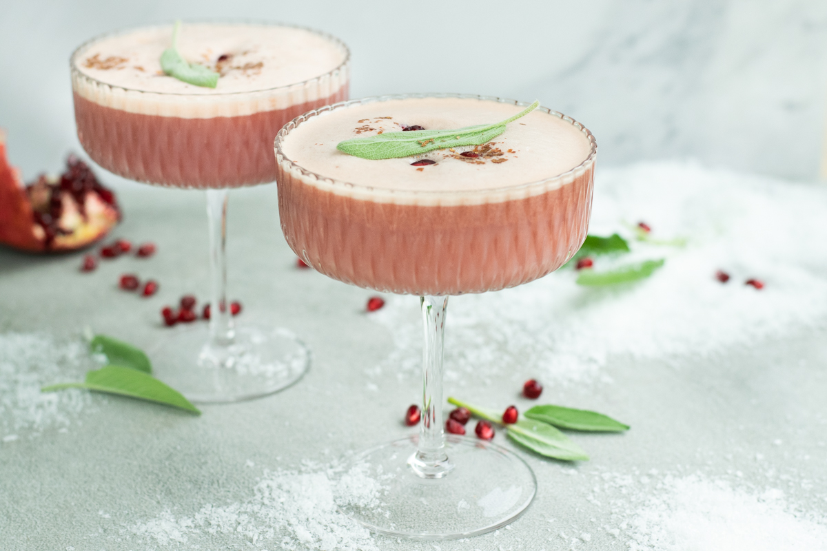 Een champagnecoupe met een zachtroze mocktail met een schuimlaagje, afgewerkt met een blaadje salie en granaatappelpitjes.