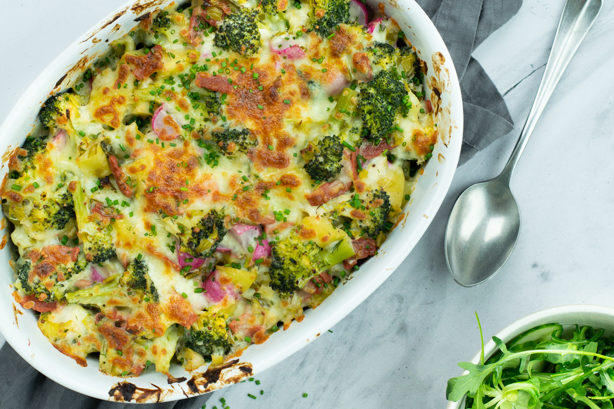 Bovenaanzicht van een geragtineerde ovenschotel met broccoli, prei, spekjes en radijsjes, op een lichte marmeren achtergrond.