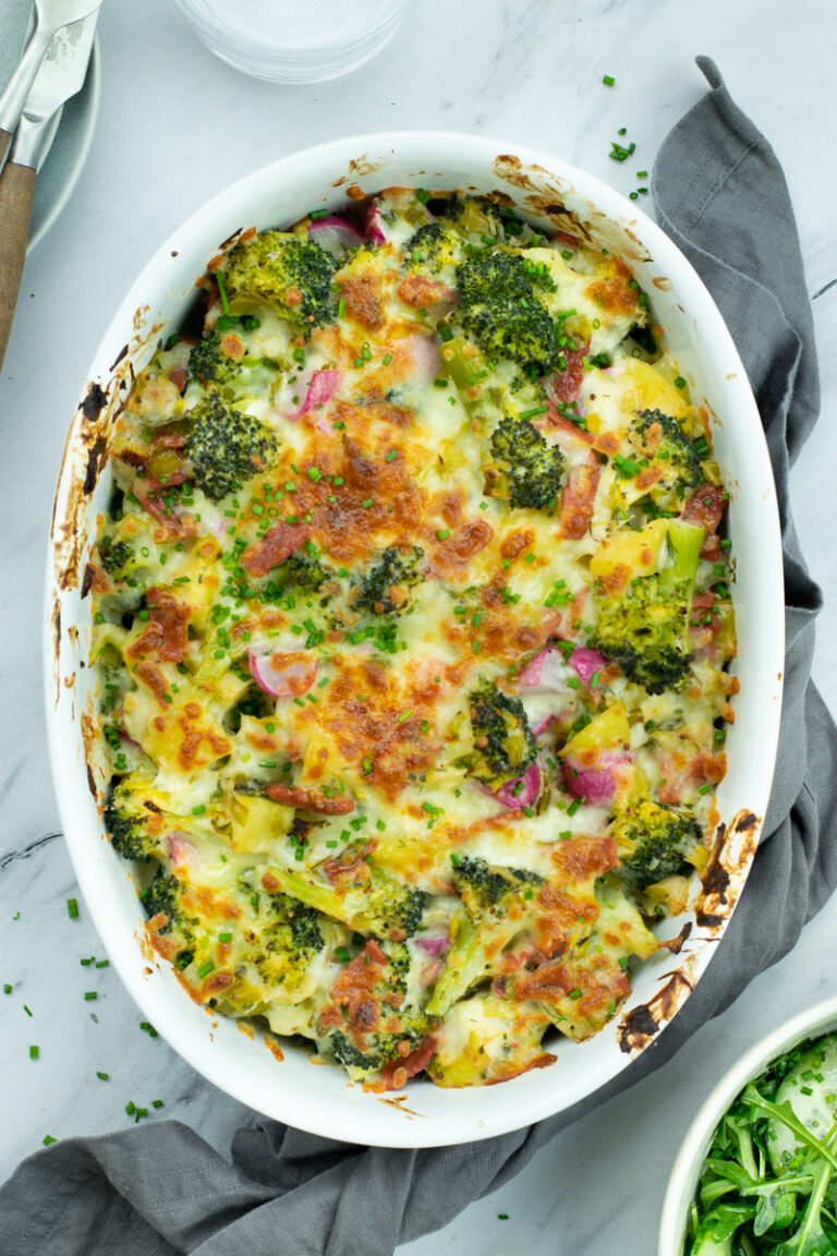 Bovenaanzicht van een geragtineerde ovenschotel met broccoli, prei, spekjes en radijsjes, op een lichte marmeren achtergrond.