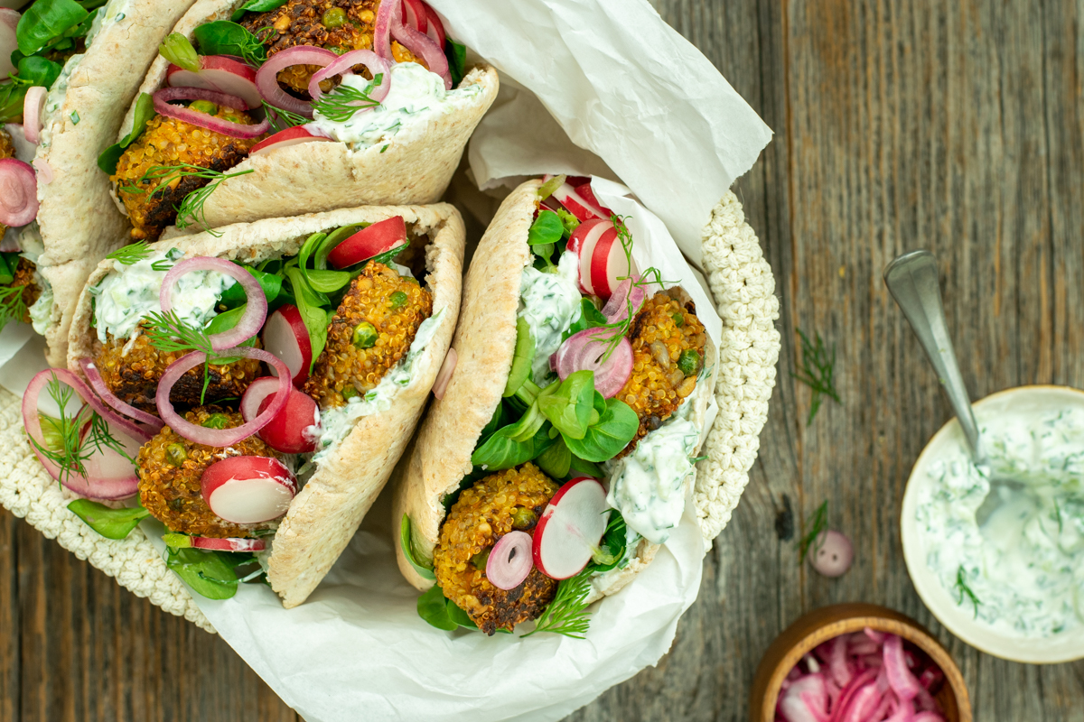 Bovenaanzicht van een mandje met 4 pitabroodjes, gevuld met wortel-quinoaburgertjes, tzatziki, radijs, veldsla en opgelegde rode ui; op een ruwe houten ondergrond.