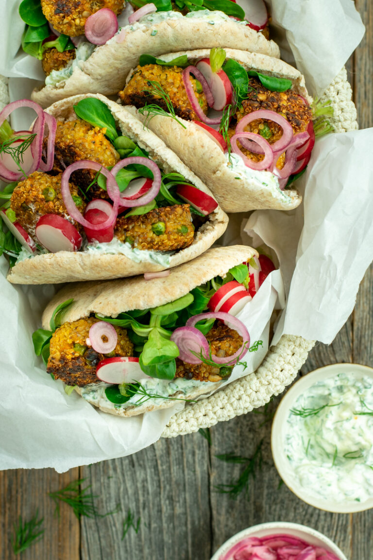 Bovenaanzicht van een mandje met 4 pitabroodjes, gevuld met wortel-quinoaburgertjes, tzatziki, radijs, veldsla en opgelegde rode ui; op een ruwe houten ondergrond.