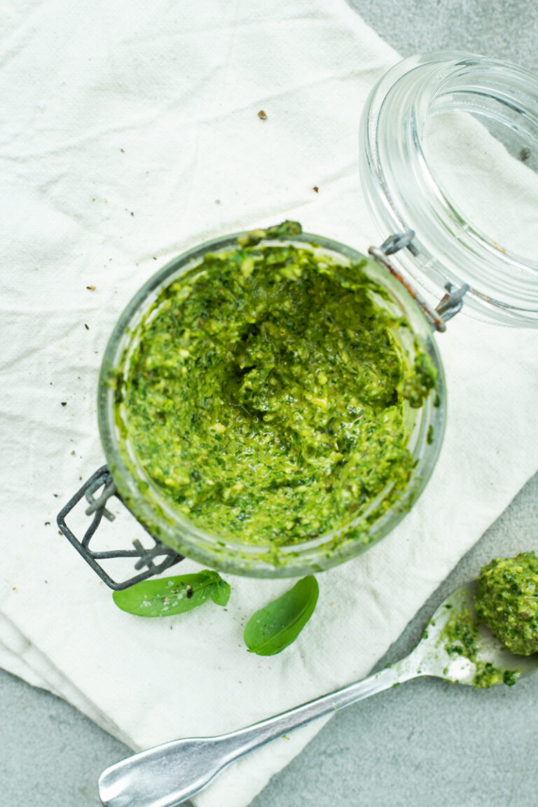 Bovenaanzicht van een bokaaltje pesto op een stoffen witte servet, op een lichtgrijze stenen ondergrond.