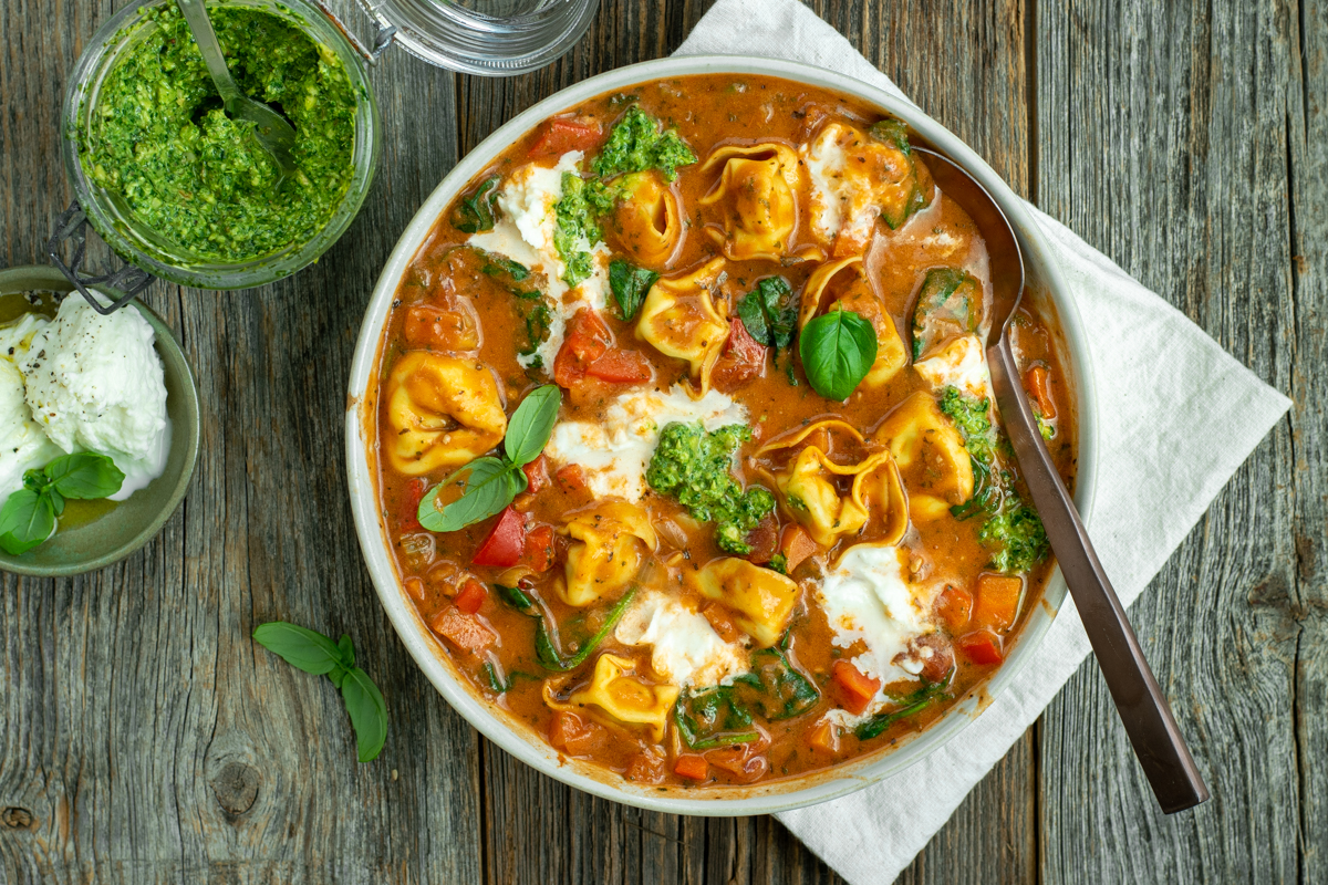 Bovenaanzicht van een diep bord met tomaten-tortellinisoep met buffelmozzarella en toefjes pesto, op een houten ondergrond.
