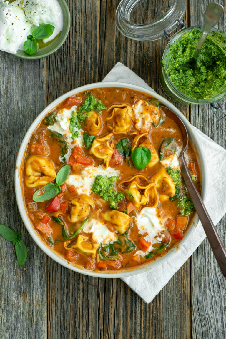 Bovenaanzicht van een diep bord met tomaten-tortellinisoep met buffelmozzarella en toefjes pesto, op een houten ondergrond.