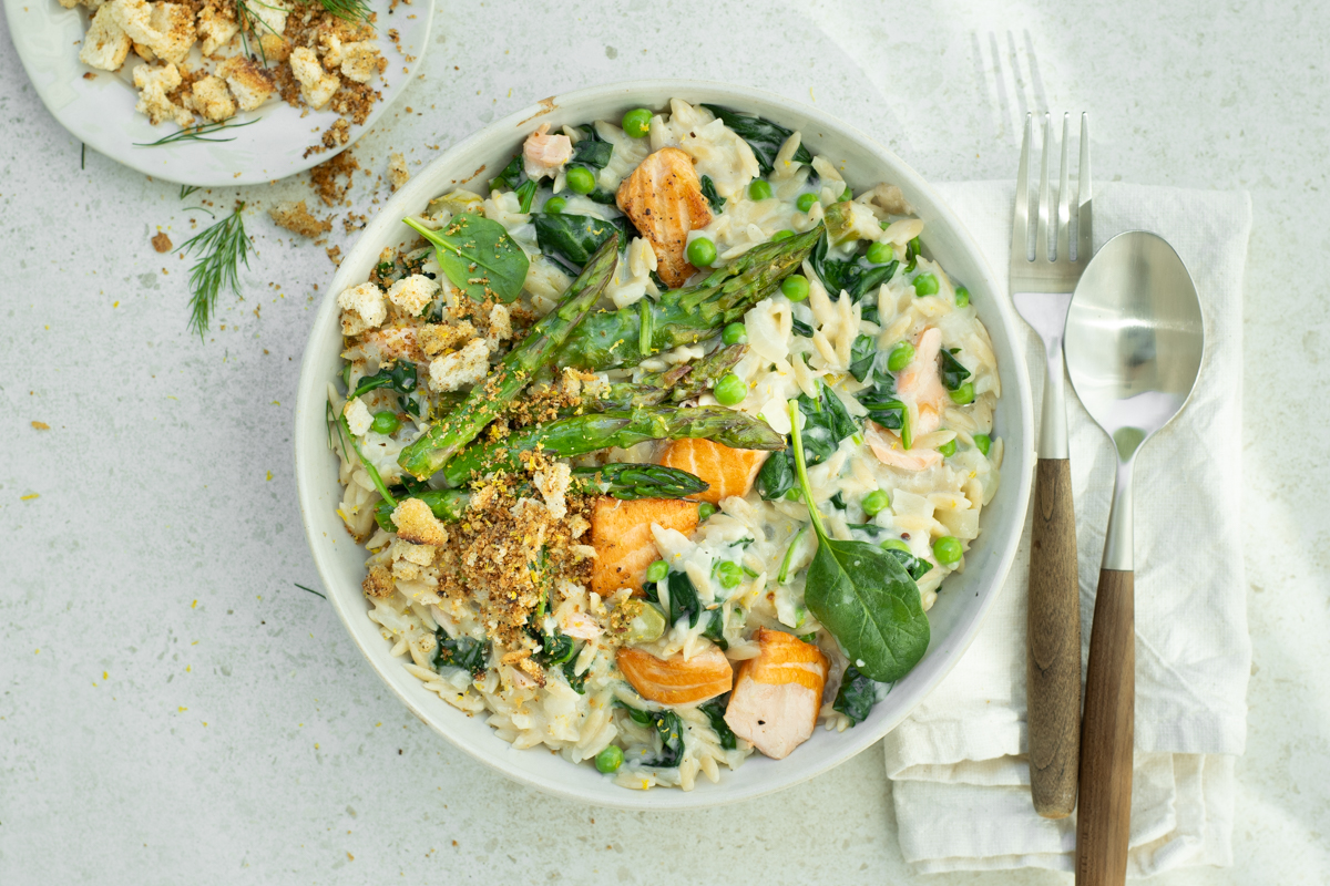 Bovenaanzicht van een wit bord, gevuld met orzotto met zalm, asperges en spinazie, op een lichtbeige achtergrond.
