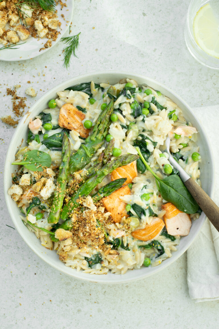 Bovenaanzicht van een wit bord, gevuld met orzotto met zalm, asperges en spinazie, op een lichtbeige achtergrond.