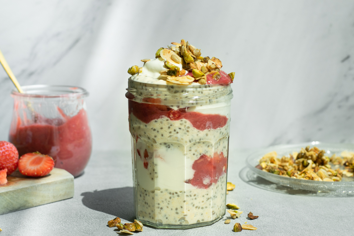 Een bokaaltje gevuld met laagjes overnight oats met chiazaad, yoghurt en roze rabarbercompote, afgewerkt met amandelschilfers en pistachenoten.