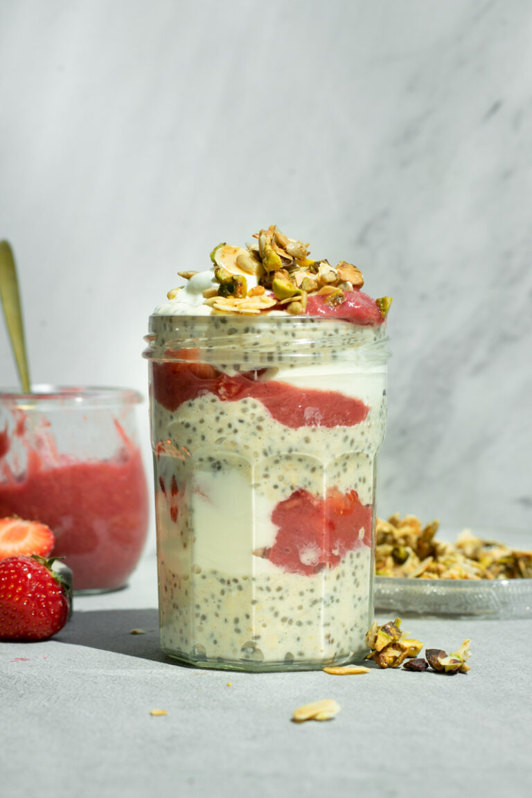 Een bokaaltje gevuld met laagjes overnight oats met chiazaad, yoghurt en roze rabarbercompote, afgewerkt met amandelschilfers en pistachenoten.