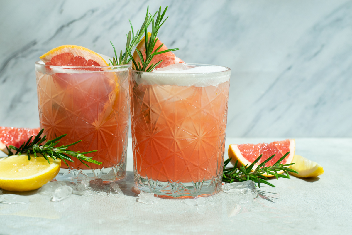 Zijaanzicht van een Fizzy pompelmoes Spritz mocktail, met een oranjeroze kleur en schuimlaagje, gedecoreerd met een schijfje pompelmoes en takje rozemarijn.