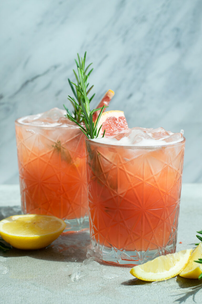 Zijaanzicht van een Fizzy pompelmoes Spritz mocktail, met een oranjeroze kleur en schuimlaagje, gedecoreerd met een schijfje pompelmoes en takje rozemarijn.