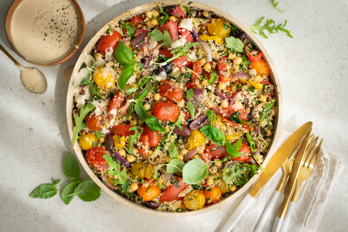 Bovenaanzicht van een ronde schaal gevuld met geroosterde tomaten, couscous, olijven, kikkererwten, afgewerkt met vers basilicum, rucola en een tahinsausje.