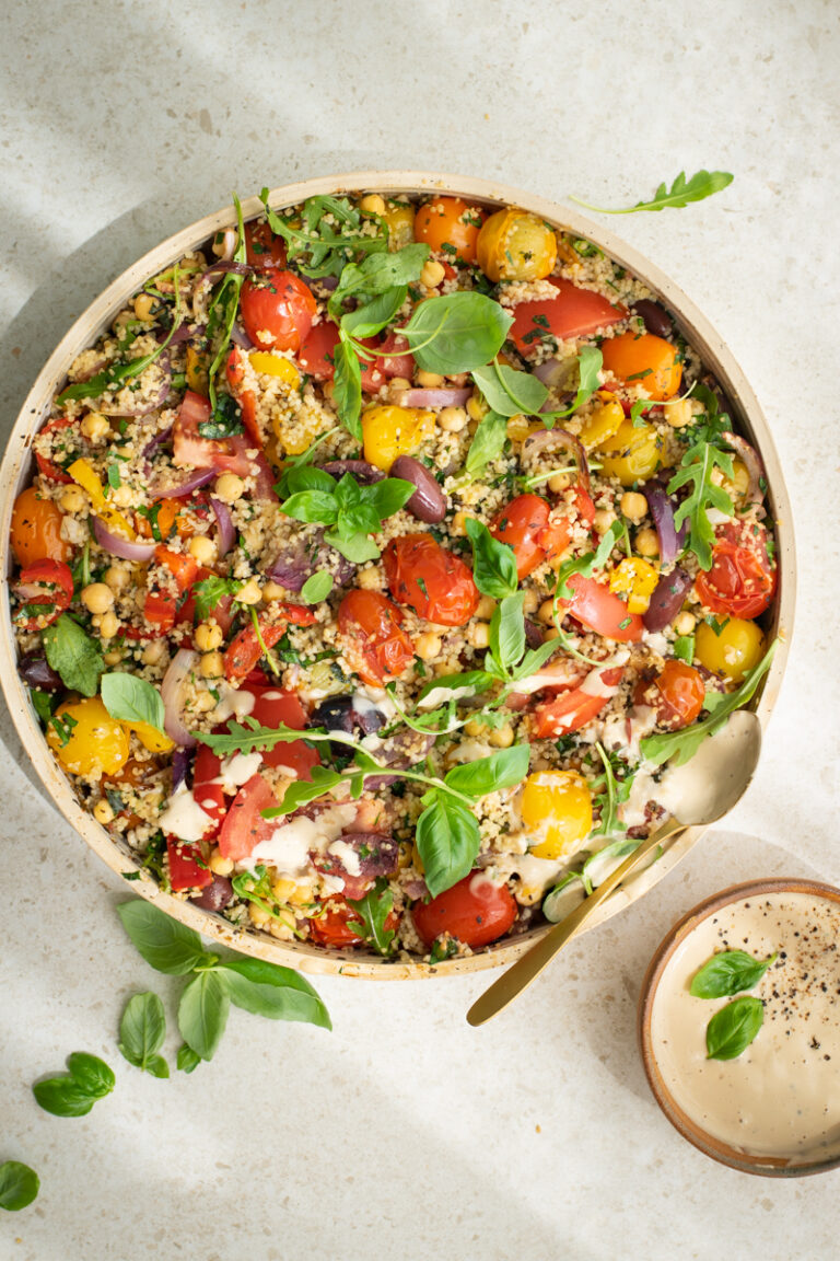Bovenaanzicht van een ronde schaal gevuld met geroosterde tomaten, couscous, olijven, kikkererwten, afgewerkt met vers basilicum, rucola en een tahinsausje.