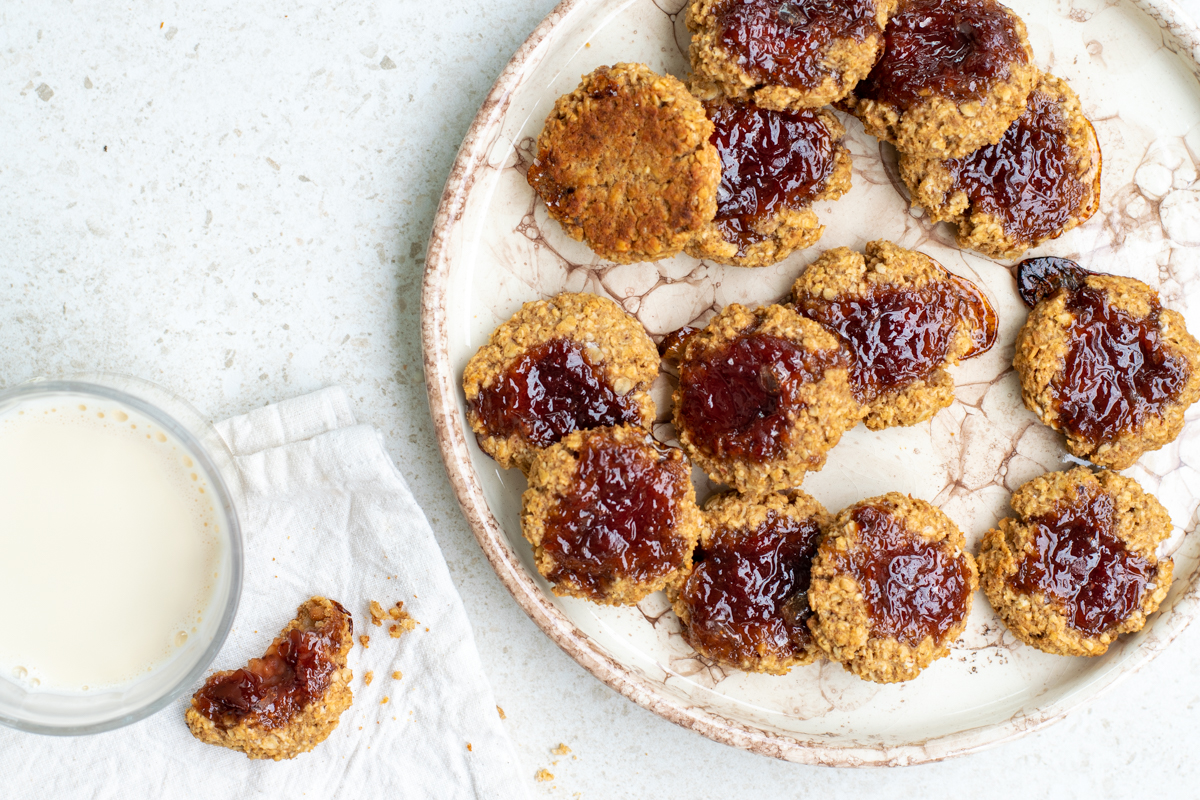 Bovenaanzicht van een beige bord vol met Havermoutkoekjes met pindakaas en aardbeienconfituur, op een beige achtergrond.